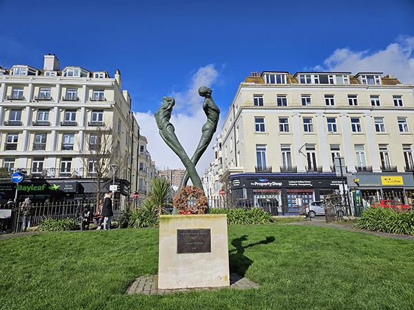 Metal sculpture of two figures looking skyward, on a plinth, in a park setting. 