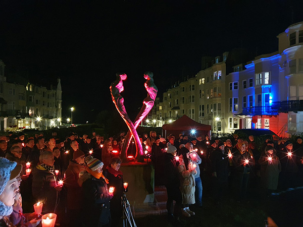 Metal sculpture of two figures looking skyward, on a plinth, lit up at night, surrounded by people holding candles