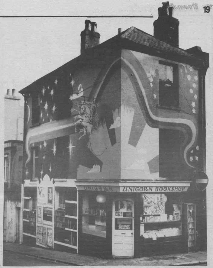 Black and white photo of a corner building with a mural of a rising sun and a unicorn. Sign above the door reads: Unicorn Bookshop